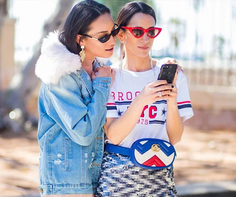 Two stylish women outdoors, one in a denim jacket and the other with a red Gucci belt bag, looking at a smartphone.