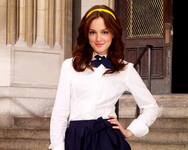 Young woman with brown hair in a white blouse and navy skirt, standing on stone steps with a confident smile.