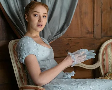 Daphne Bridgerton sitting on a chair in a Regency-style dress, holding sheer gloves, against a wooden paneled background.