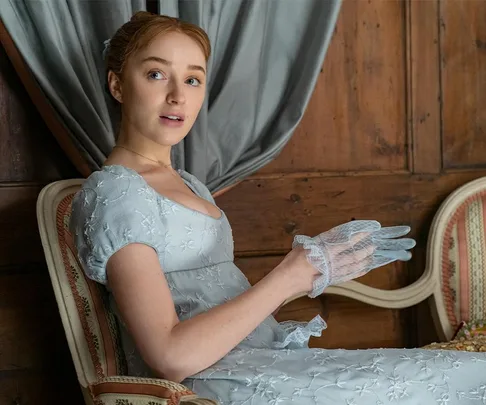 Daphne Bridgerton sitting on a chair in a Regency-style dress, holding sheer gloves, against a wooden paneled background.