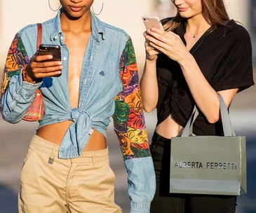 Two women walking, using smartphones; colorful shirt and Alberta Ferretti bag.