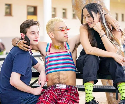 Three friends sit together on a bench; one in a striped top with blonde hair, another with long dark hair, and one holding a phone.