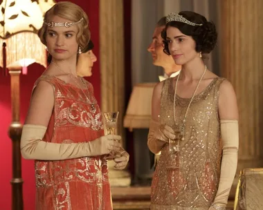 Two women in elegant 1920s dresses and tiaras holding champagne glasses at a glamorous event.