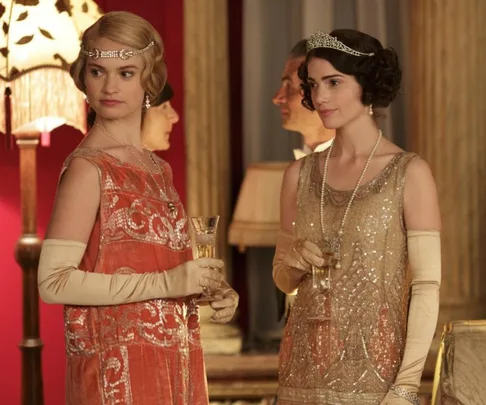 Two women in elegant 1920s dresses and tiaras holding champagne glasses at a glamorous event.