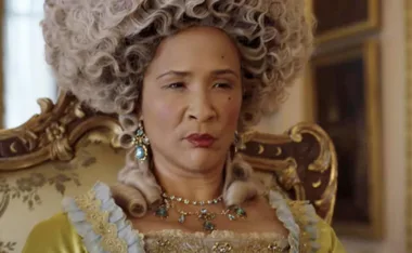 Woman dressed as Queen Charlotte in ornate gown and wig from the series "Bridgerton," seated in a lavish room.