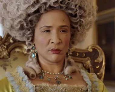 Woman dressed as Queen Charlotte in ornate gown and wig from the series "Bridgerton," seated in a lavish room.