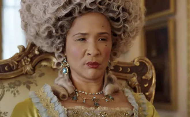 Woman dressed as Queen Charlotte in ornate gown and wig from the series "Bridgerton," seated in a lavish room.