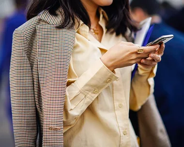 Woman in a yellow shirt and plaid coat checks her phone while holding a book, seen from chest up.