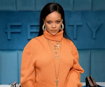 Rihanna in an orange sweater with gold jewelry, standing in front of a blue Fenty sign.