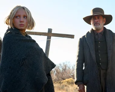 A young girl and an older man in period clothing stand outdoors under a blue sky in a rural setting.