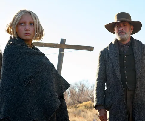 A young girl and an older man in period clothing stand outdoors under a blue sky in a rural setting.