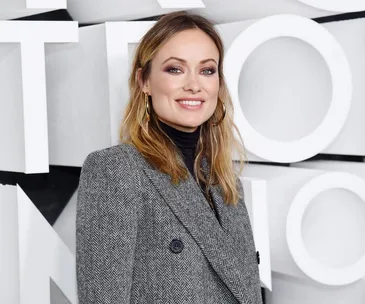 A person smiling, wearing a gray herringbone coat, standing in front of a geometric wall background.