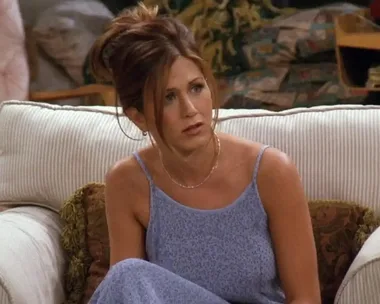 Rachel Green from Friends, in a blue dress, sitting on a beige couch with a thoughtful expression.