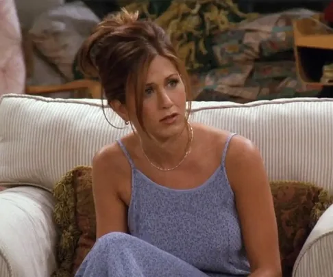 Rachel Green from Friends, in a blue dress, sitting on a beige couch with a thoughtful expression.