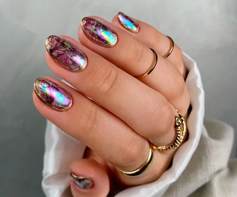 A hand with rainbow opal nails, shiny and colorful, adorned with gold rings.