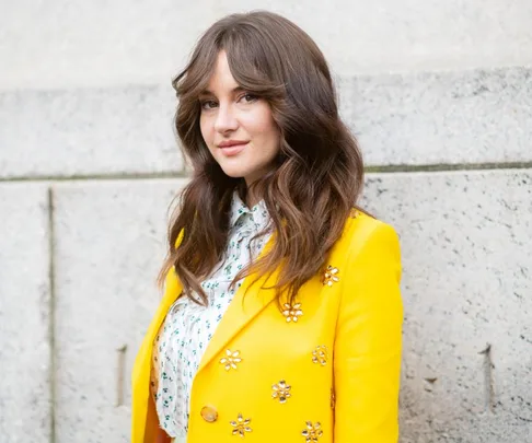 Shailene Woodley in a yellow embellished blazer, standing against a stone wall, looking at the camera.