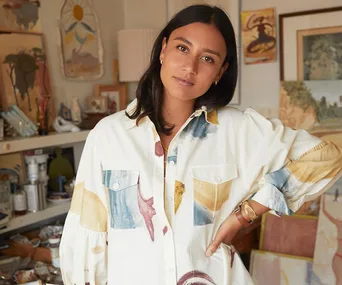 Woman in a colorful shirt stands in an art-filled studio, looking at the camera with a relaxed expression.