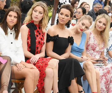 Five women seated front row at a fashion event, dressed in stylish and elegant outfits, watching the show.