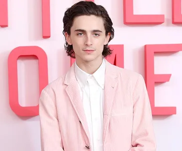 Man in a pink suit stands in front of a pink and white background, looking directly at the camera.