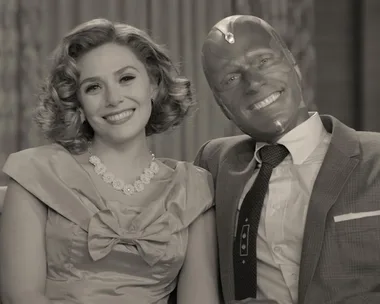 A couple, a woman in a 1950s dress and a man in a suit with a robotic face, sit smiling on a couch in a black-and-white scene.