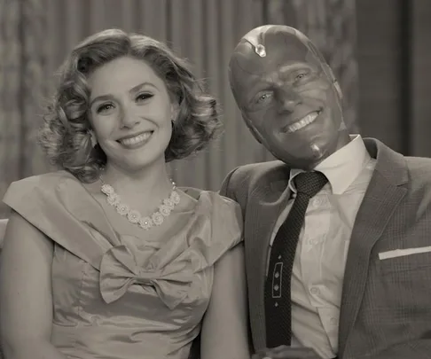 A couple, a woman in a 1950s dress and a man in a suit with a robotic face, sit smiling on a couch in a black-and-white scene.