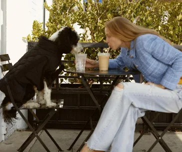 Woman and dog in a coat share drinks at outdoor cafe table.