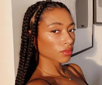 A woman with glowing skin and braided hair looks toward the camera in soft lighting.