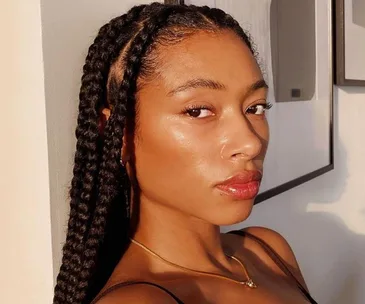 A woman with glowing skin and braided hair looks toward the camera in soft lighting.