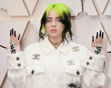 Person with black and neon green hair wearing a white jacket, showcasing intricate rings and long black nails.