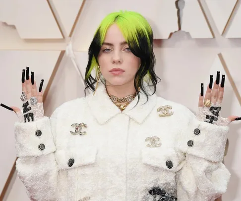 Person with black and neon green hair wearing a white jacket, showcasing intricate rings and long black nails.