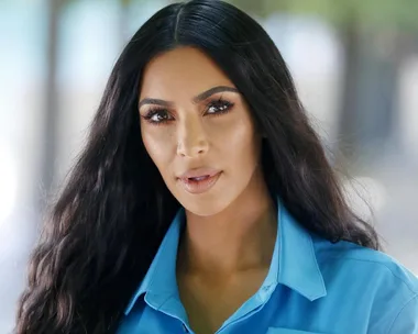 Celebrity with long dark hair and bold eyebrows, wearing a blue shirt, looking directly at the camera.