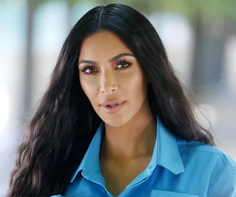 Celebrity with long dark hair and bold eyebrows, wearing a blue shirt, looking directly at the camera.