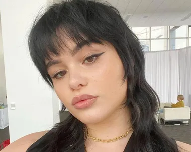 Woman with black mullet hairstyle, eyeliner, gold chain, in a brightly lit room with white curtains in the background.