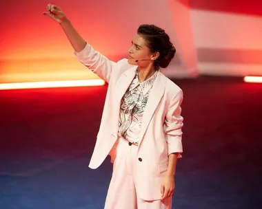 A woman in a white suit with a headset gestures on stage with red lighting in the background.