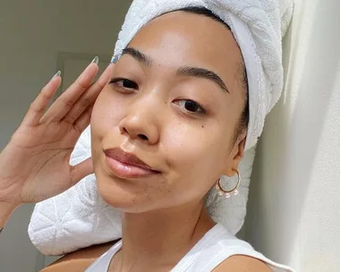 Woman with natural skin, wearing a towel on her head, poses with minimal makeup and a slight smile.