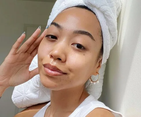 Woman with natural skin, wearing a towel on her head, poses with minimal makeup and a slight smile.