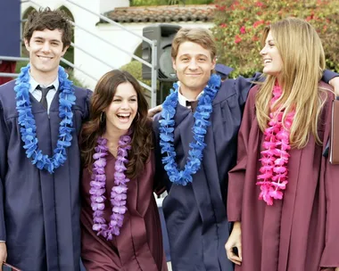 Four people in graduation gowns and leis smile together at an outdoor ceremony.