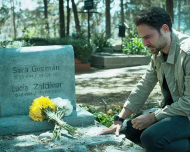 A man kneels by a gravestone for Sara Guzman and Lucía Zaldivar, with flowers placed on it, in a wooded cemetery.