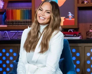 A woman with long hair in a white outfit smiles, seated in a studio with colorful background elements and books.