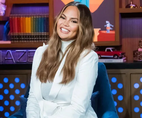 A woman with long hair in a white outfit smiles, seated in a studio with colorful background elements and books.