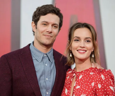 A smiling couple poses together, with the woman in a red patterned dress and the man in a maroon jacket and blue shirt.