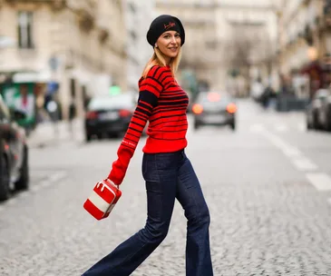 Woman in a red striped sweater and beret, carrying a small bag, smiles and walks on a cobblestone street in Paris.