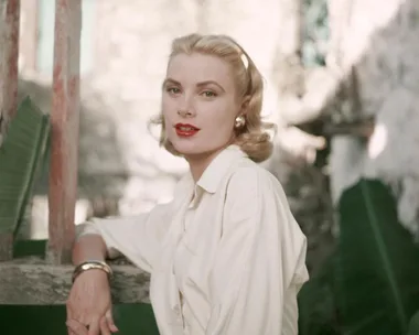 A woman with blonde hair and red lipstick wearing a white shirt, posed outdoors near greenery.