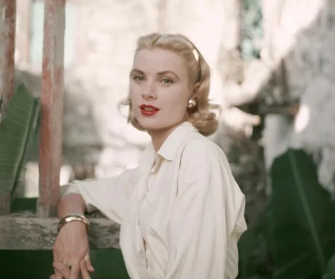 A woman with blonde hair and red lipstick wearing a white shirt, posed outdoors near greenery.