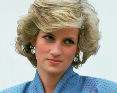 A person with styled, voluminous blonde bob, wearing diamond heart earrings and a blue patterned blouse.