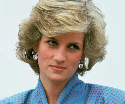 A person with styled, voluminous blonde bob, wearing diamond heart earrings and a blue patterned blouse.