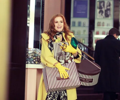 A woman in a stylish yellow coat holds shopping bags from Henri Bendel, smiling inside a store.