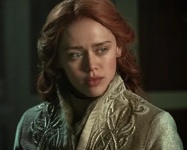 A woman with red hair gazes intently to the side, wearing an ornate embroidered coat, in a dimly lit setting.