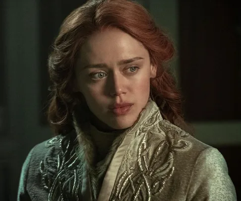 A woman with red hair gazes intently to the side, wearing an ornate embroidered coat, in a dimly lit setting.