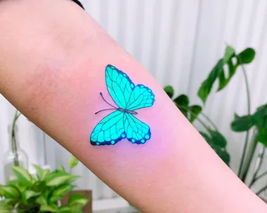 A glowing blue butterfly tattoo on a person's arm, set against a blurred plant background, showcasing UV ink.